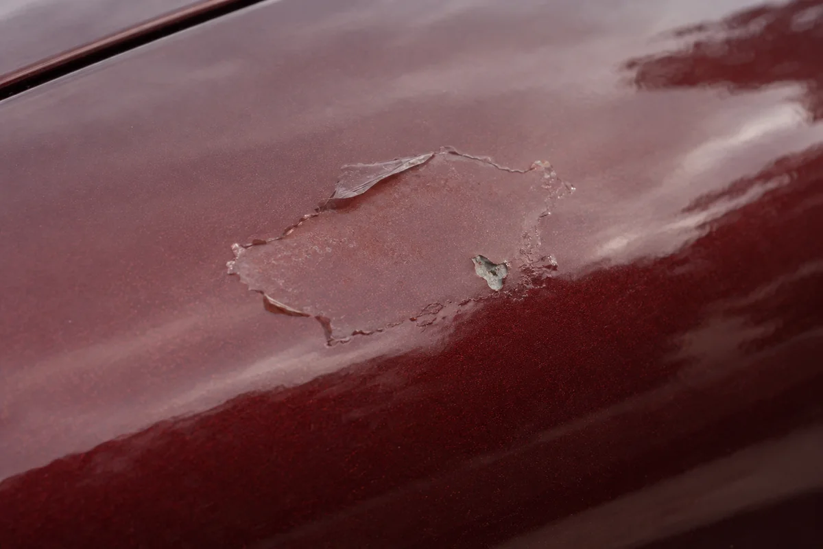 A small patch of lifting clearcoat and a stone chip on burgundy paint shows raised edges that need assessment before wrapping